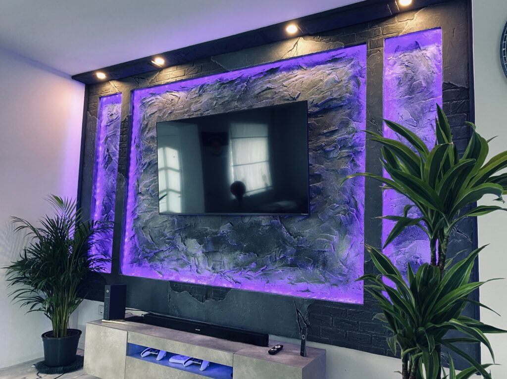 Moderne TV-Akzentwand aus dunklem Kunststein mit markanter Felsstruktur, umrahmt von violetter LED-Hintergrundbeleuchtung und Deckenstrahlern.