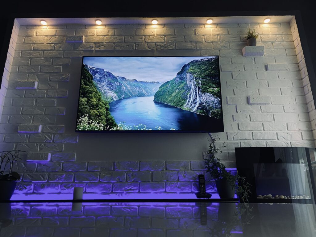 Wandmontierter Fernseher an einer hellen Kunststein-Wand in Backsteinstruktur mit Deckenstrahlern und violetter LED-Bodenbeleuchtung.