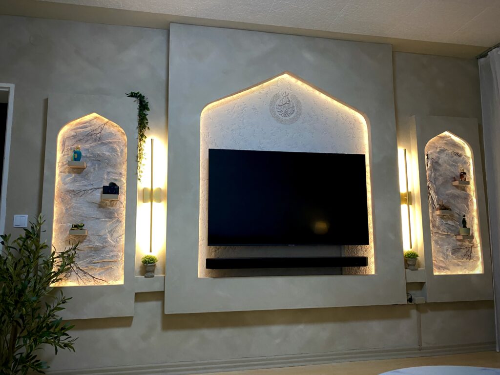 Drei vorgesetzte Wandelemente mit orientalischen Spitzbögen, warmen LED-Lichtleisten, Wandleuchten und Nischen für Dekoration um den Fernseher.