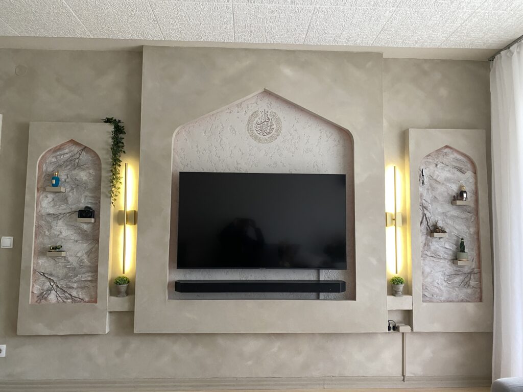 Fertige helle TV-Wand mit zentralem Bogen für den Fernseher, seitlichen Nischen mit orientalischem Relief und Wandleuchten bei Tageslicht.