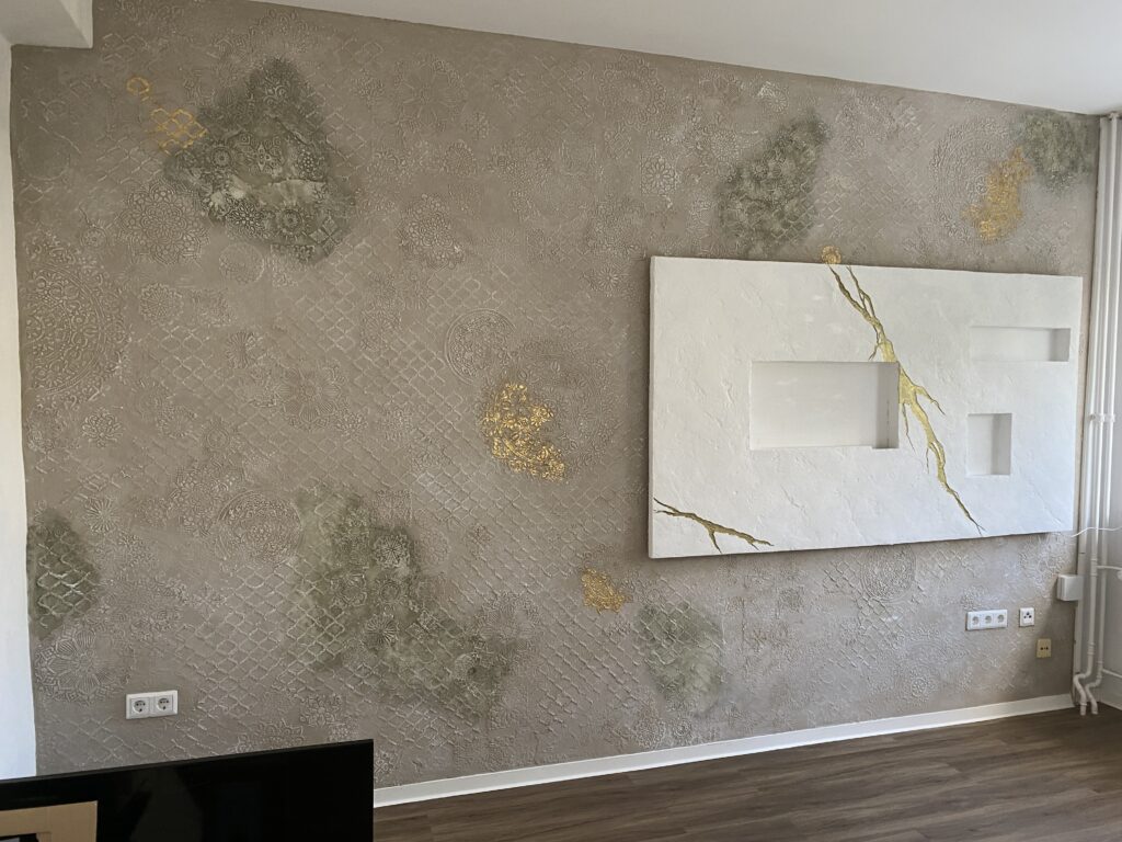 Vorgesetzte weiße TV-Wand aus Kunststein mit goldener Bruchlinie und Nischen vor einer beigen Akzentwand mit floralen und orientalischen Reliefs.