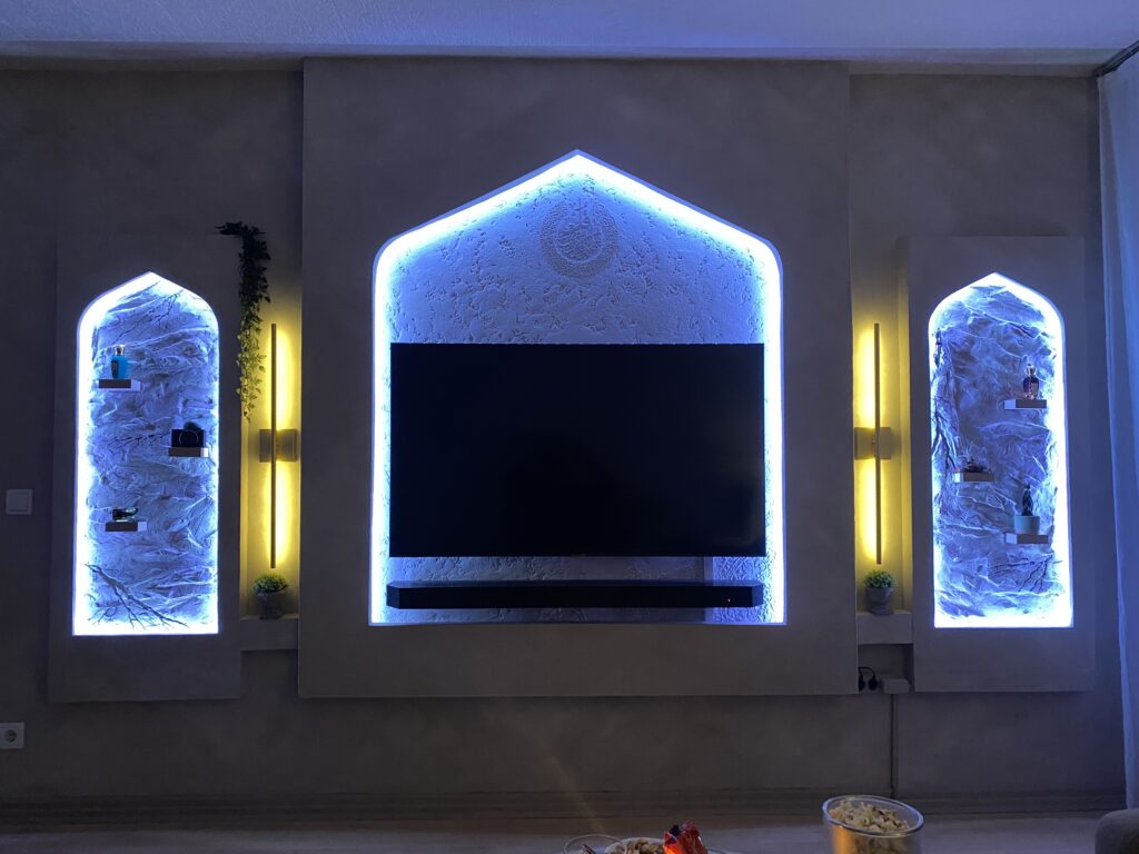 TV-Wand im abgedunkelten Raum mit leuchtend blauer LED-Hintergrundbeleuchtung entlang der Bogenkonturen und warmen Seitenleuchten.