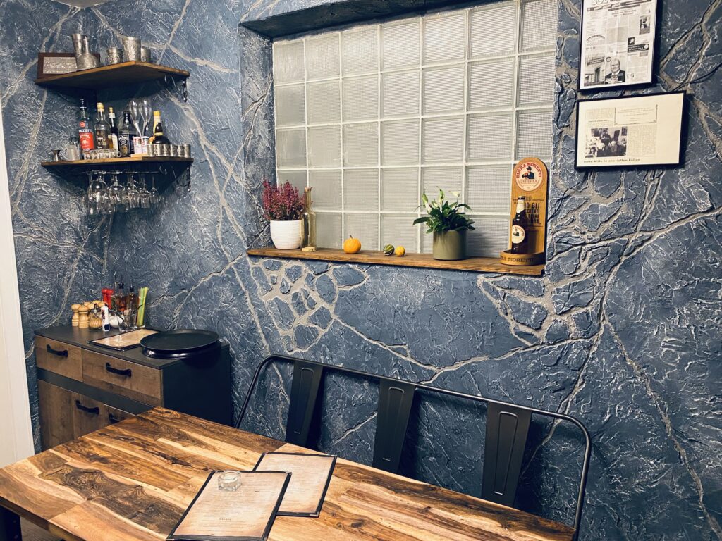 Rückwand in einem Restaurant aus blaugrauem Kunststein mit ausgeprägter Felsstruktur und Aderung, Glasbaustein-Fenster sowie rustikalen Holzmöbeln.
