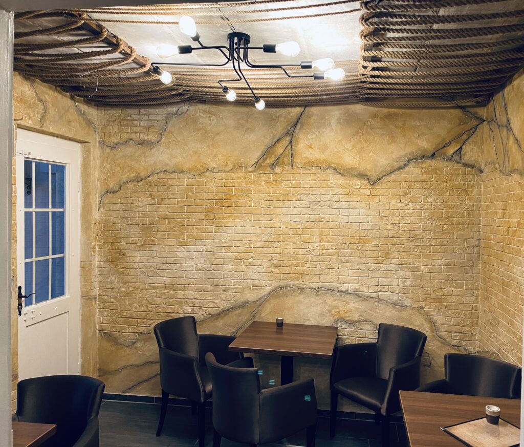 Gastronomiebereich mit warmgelber Kunststein-Wand in Sandstein- und Backsteinoptik, holzverzierte Seildecke, Tische und Lederstühle.