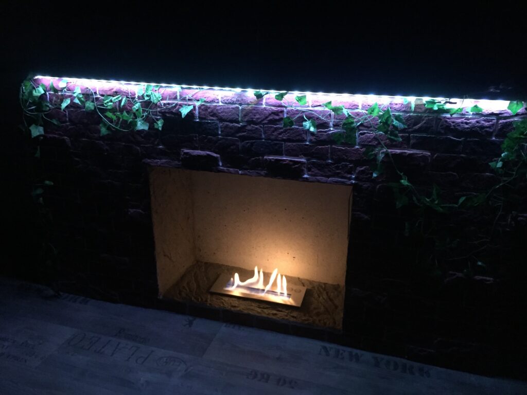 Nachgerüsteter künstlicher Kaminsims aus dunklem Kunststein mit brennendem Bioethanol-Kamin, Efeudekoration und LED-Lichtleiste.