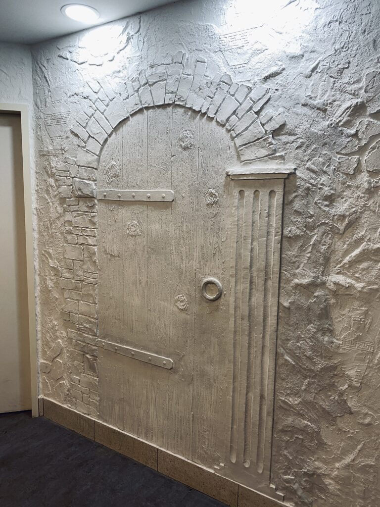 Weißes Kunststein-Wandrelief im Sanitärbereich mit eingearbeiteten griechischen Krügen, einer rustikalen Pforte und antiken Säulenelementen.