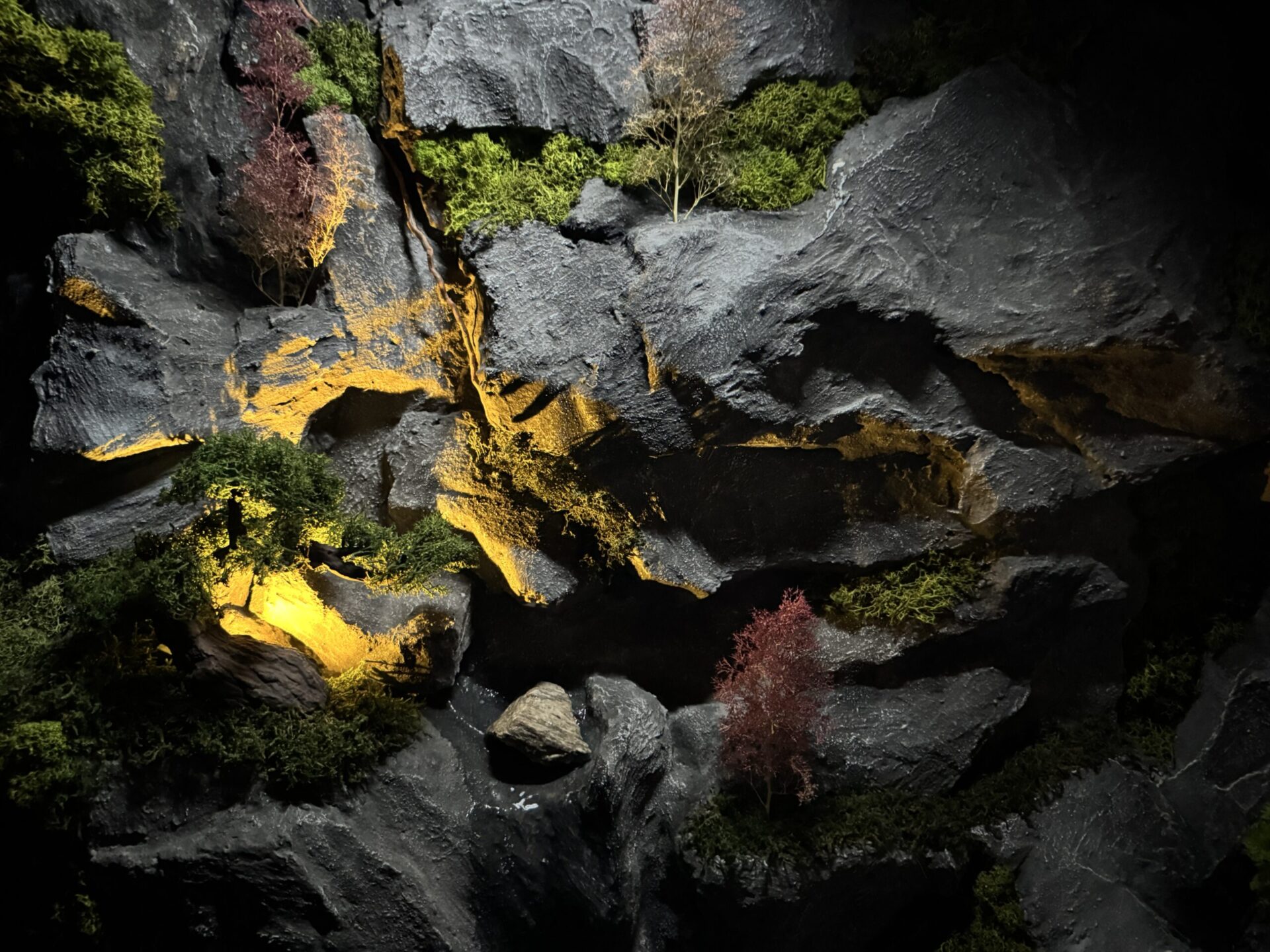 Detailansicht einer Kunststein-Wasserfallwand mit rauer Gesteinsstruktur, Moos, Pflanzendetails und warmen Lichtakzenten.