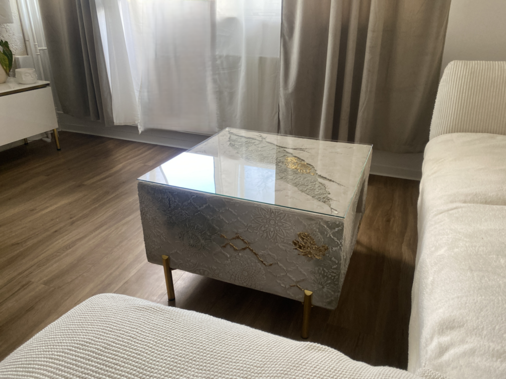 Quadratischer Couchtisch mit eingeprägten Spitzen- und Mandala-Reliefs in Beigetönen, Blattgold-Akzenten, messingschimmernden Beinen und einer Glasplatte.
