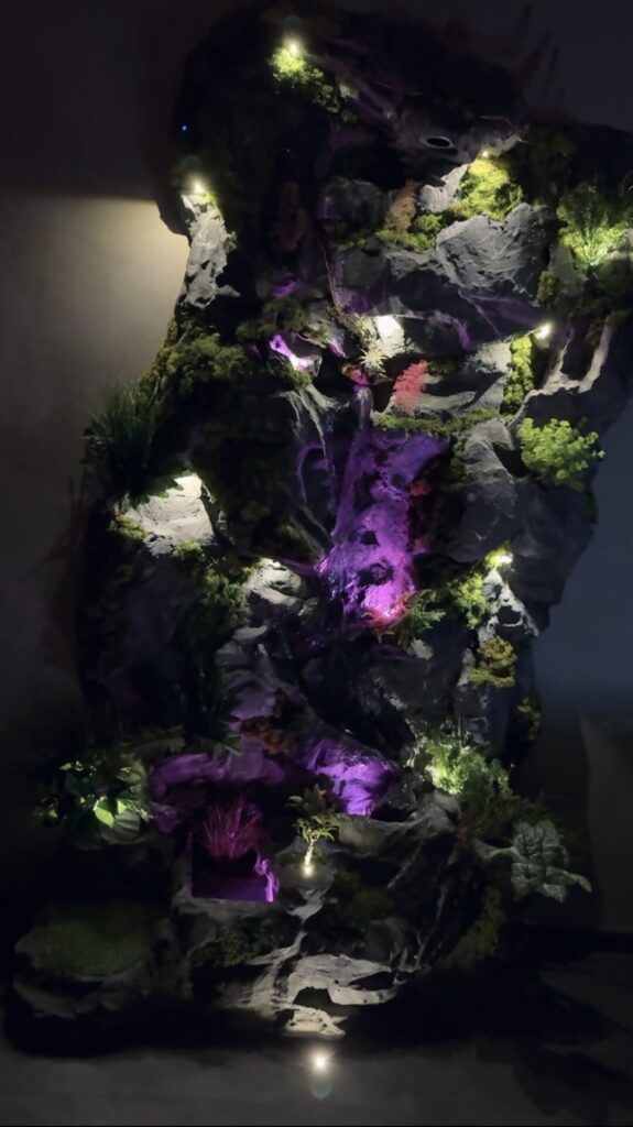 Künstliche Wasserfallwand in Felsoptik mit atmosphärischer violetter und grüner LED-Beleuchtung sowie Pflanzenelementen.